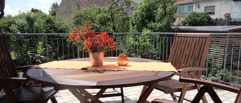 A round wooden table with four chairs, a vase with red flowers, and a candle on a balcony with a view of houses and trees.
