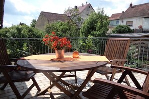 Una mesa redonda de madera con cuatro sillas, un jarrón con flores rojas y una vela en un balcón con vistas a casas y árboles.
