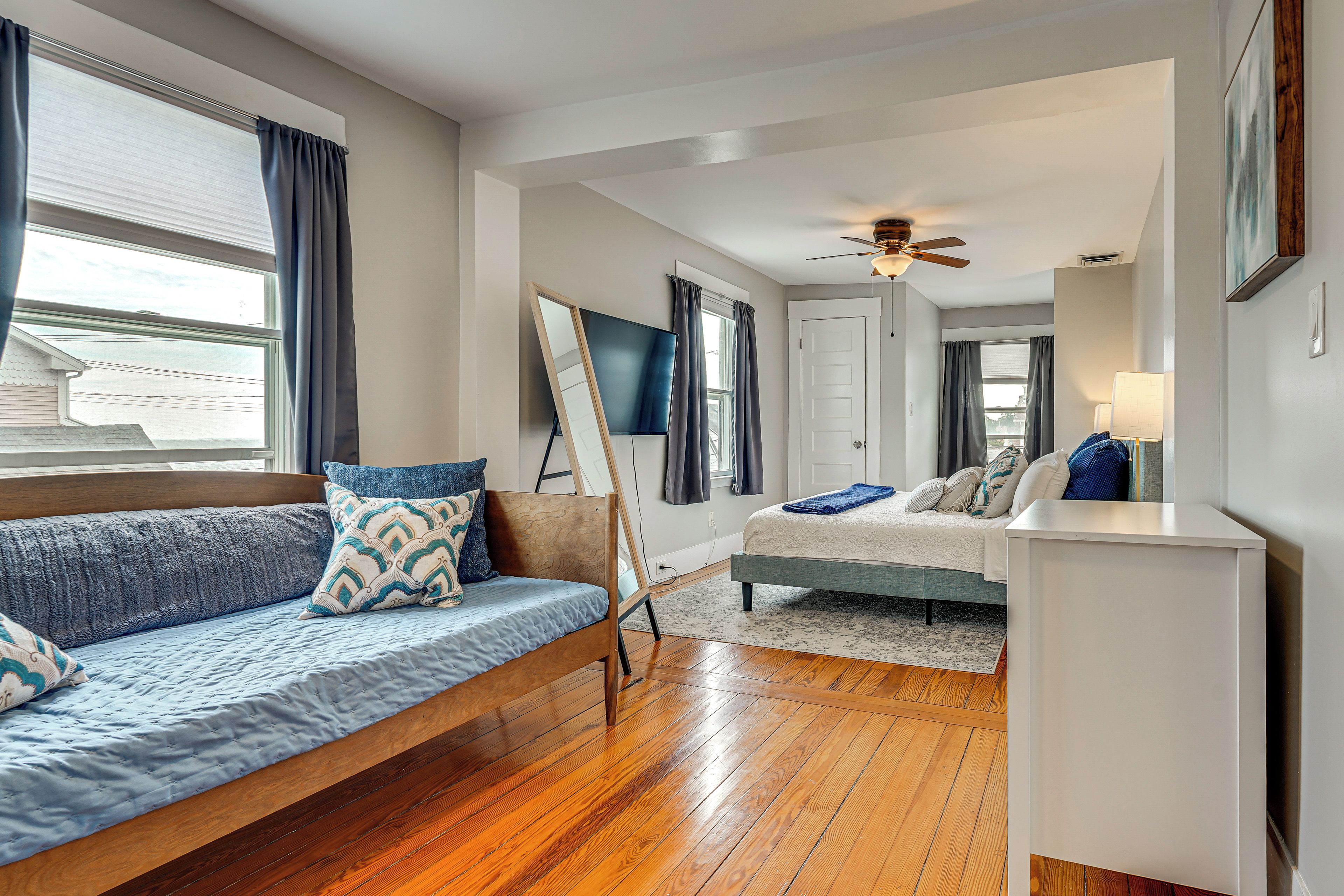 A modern bedroom with a wooden floor, a bed with a blue and white patterned bedspread, a blue sofa with matching cushions, and a ceiling fan.