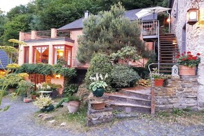Une maison à deux étages avec un escalier en pierre, un balcon et un jardin où poussent différentes espèces de plantes.