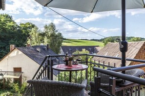 Ein Balkon mit Korbmöbeln, einem Tisch mit einer Weinflasche und Blick auf ein grünes Feld sowie Gebäude.