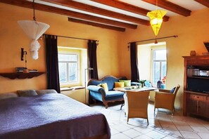 Ein Hotelzimmer mit einem Bett, einem Sofa, einem Esstisch und Stühlen. Es gibt Fenster mit Vorhängen, eine Hängelampe und ein Regal mit Büchern.