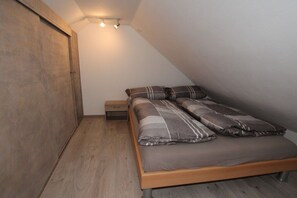 Une chambre avec un plafond en pente, un lit recouvert d’une couvre-lit à motifs gris et marron, une petite table de chevet et une armoire en bois.
