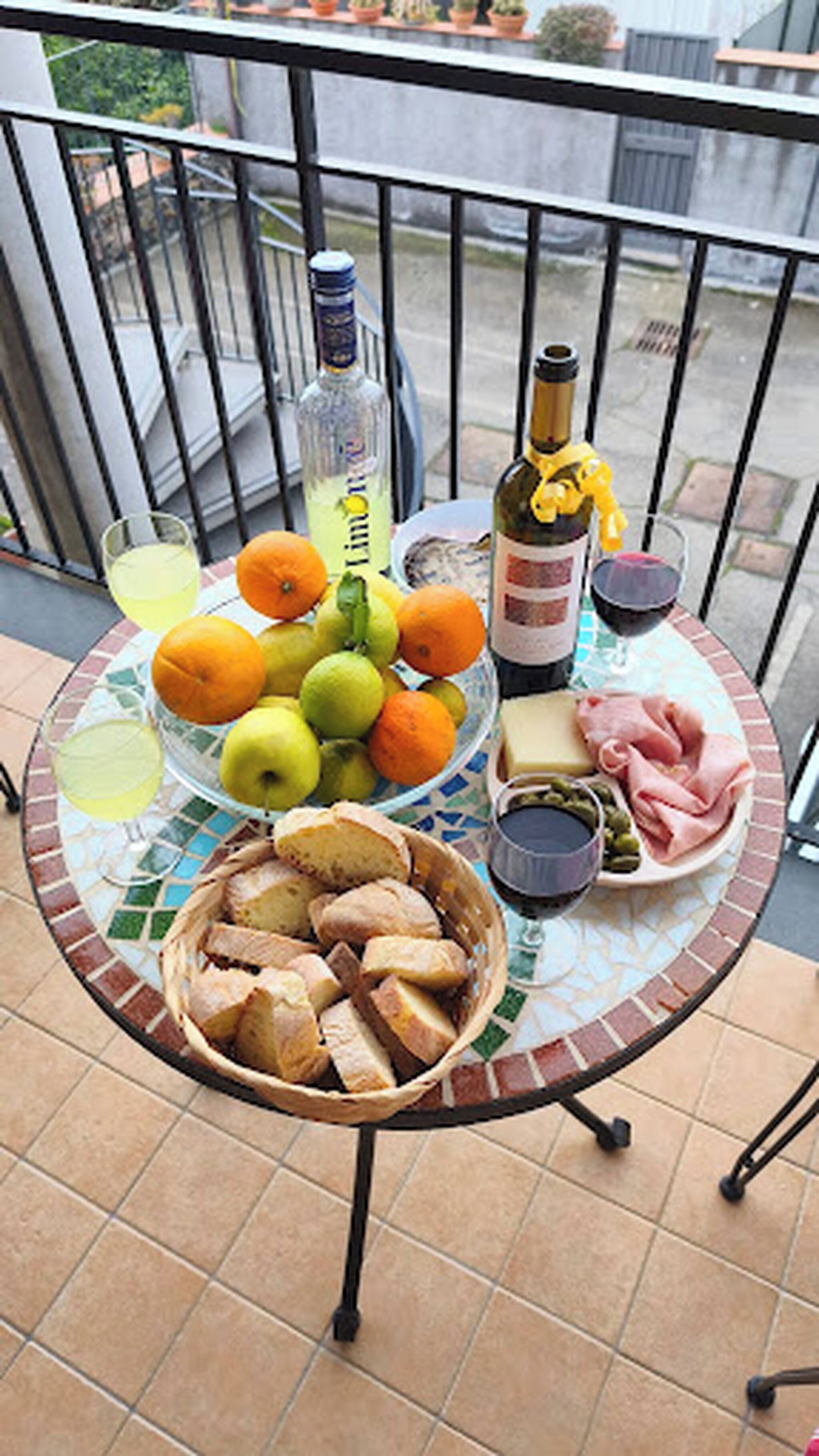 Una mesa con un frutero, botellas y una cesta de pan en un balcón.