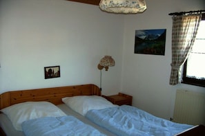 A bedroom with two single beds, a wooden headboard, a bedside table, a lamp, a framed picture, a window with curtains, and a radiator. - Holiday home Alpkönig (Missen)