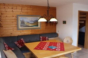 A room with a wooden wall, a sofa, a table with a red placemat, and a hanging light fixture. - Holiday home Alpkönig (Missen)