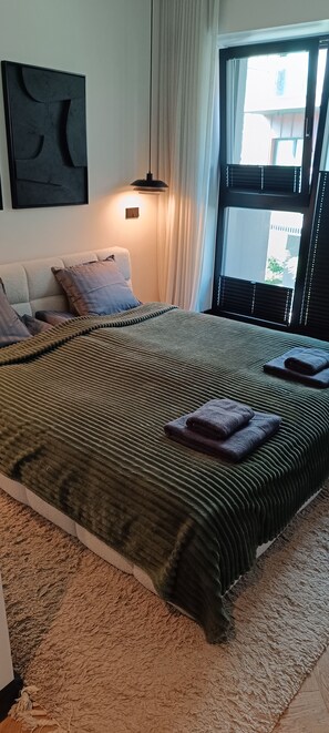 A bedroom with a large bed, a hanging light, a window with blinds, and a wall-mounted artwork. - Kalaranna family & business (Tallinn)
