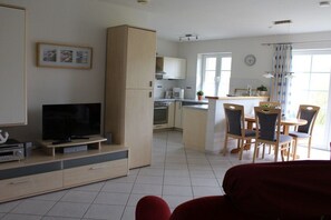 A modern living room with a flat-screen TV, a dining area with wooden chairs, and a kitchen with white cabinets. - House Swallow's Nest (Nordstrand)
