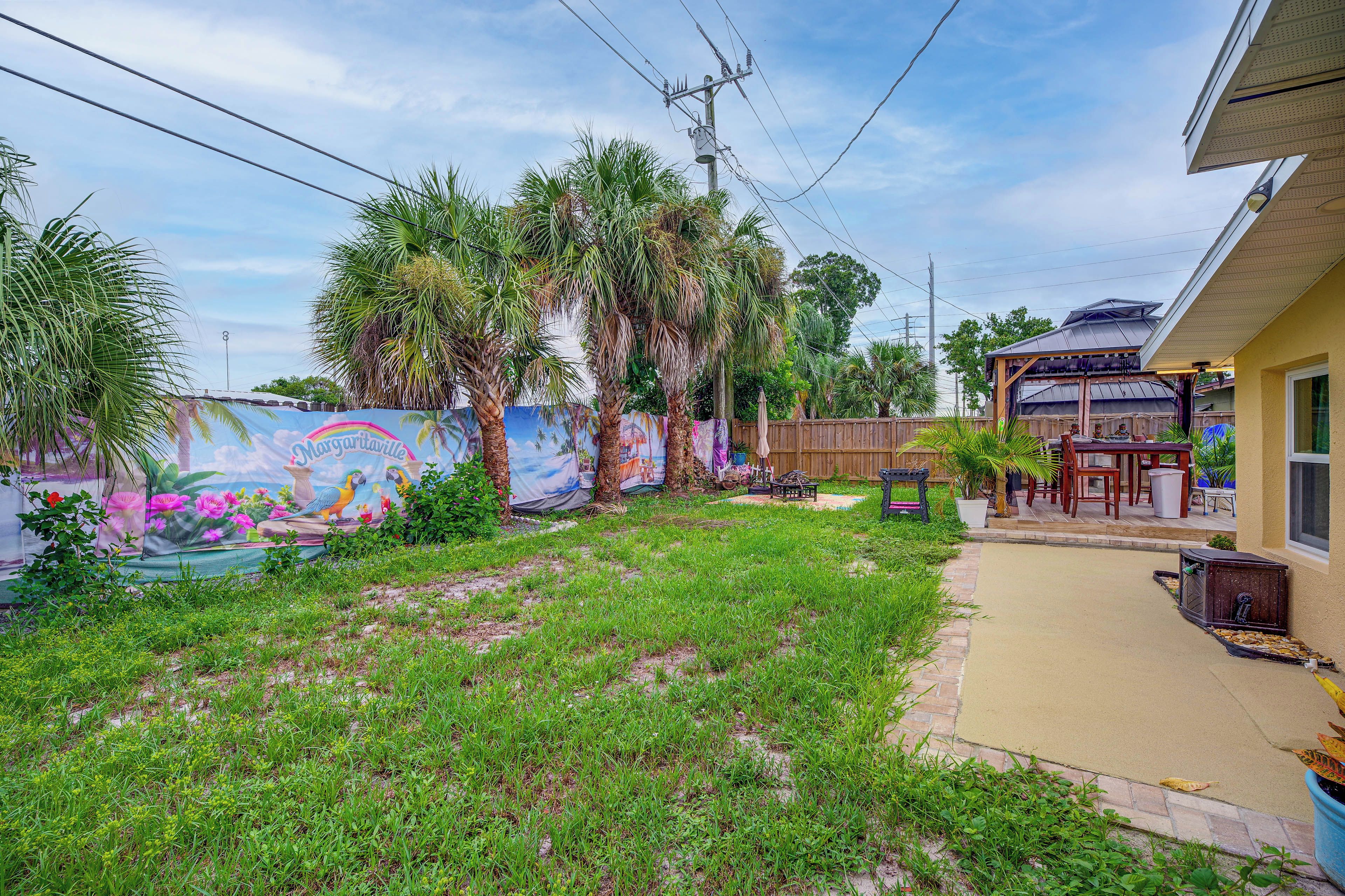 A residential backyard with a mural, palm trees, a wooden fence, and a house with a covered porch.