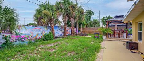 A residential backyard with a mural, palm trees, a wooden fence, and a house with a covered porch.