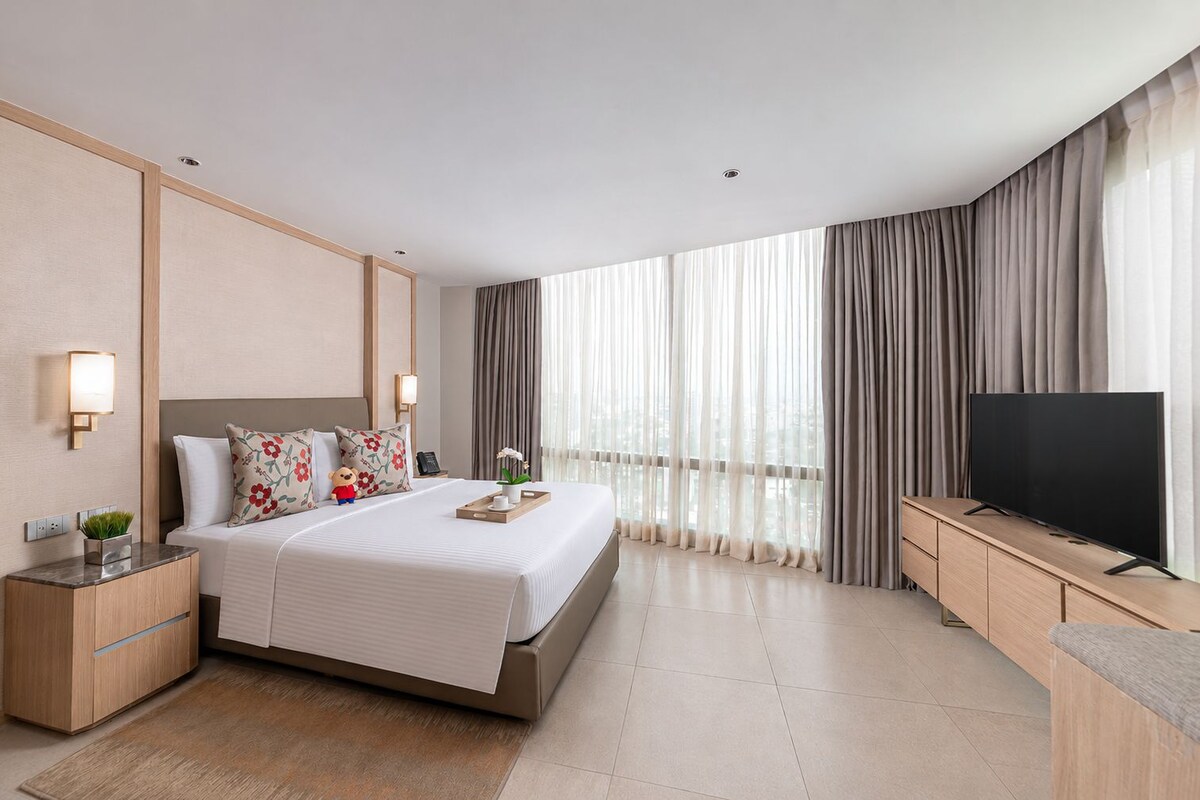 Habitación de hotel moderna con una cama grande, mesitas de noche, televisor de pantalla plana y vistas a la ciudad a través de cortinas translúcidas.