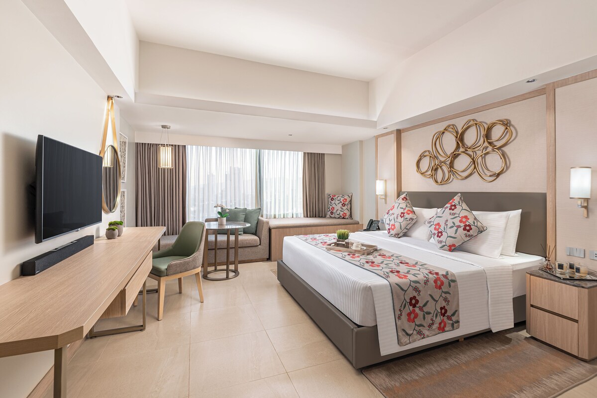 Habitación de hotel con cama, mesita de noche, escritorio, silla, televisor y vista a la ciudad por la ventana.
