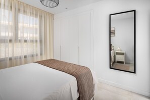 Ein Schlafzimmer mit einem Bett, einem Nachttisch mit Lampe, einem großen Spiegel und einem Fenster mit transparenten Vorhängen.