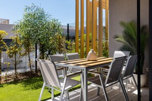 En udendørs terrasse med et træbord og stole, omgivet af grønt og en moderne bygning.