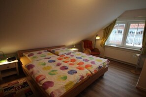 A bedroom with two beds, a chair, a lamp, a radiator, and a window with curtains. - Windloper (Wallbox!) (Norden)