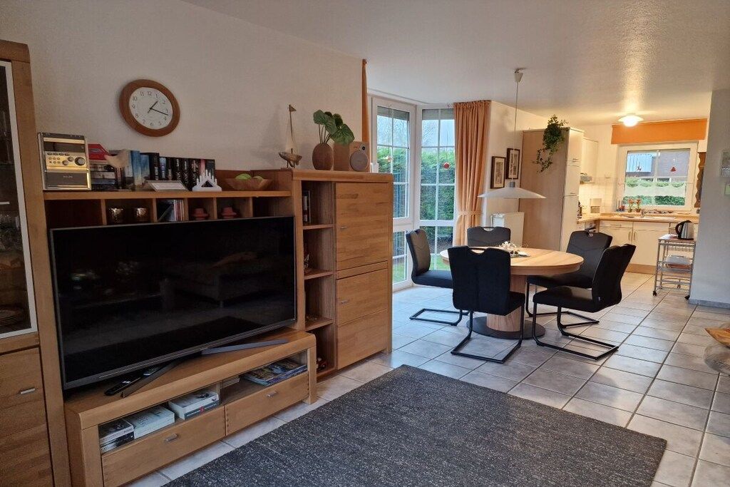 Een woonkamer met een televisie, houten meubels, een eethoek met een tafel en stoelen, en een keuken op de achtergrond.