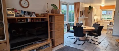 Sala de estar con televisor, muebles de madera, comedor con mesa y sillas, y cocina al fondo.