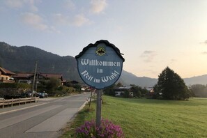 Ein Willkommen-Schild in ländlicher Umgebung, mit einer Straße, Häusern und Bergen im Hintergrund.