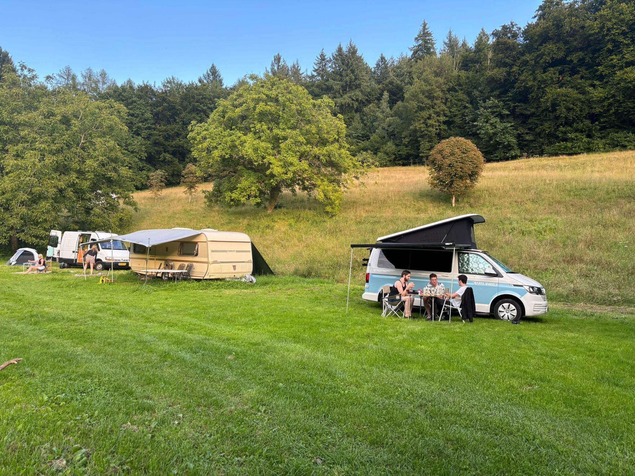 A campsite with a van and a trailer, grassy area, and trees in the background.