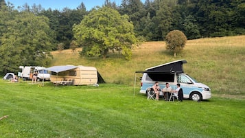 A campsite with a van and a trailer, grassy area, and trees in the background.