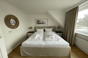 Ein Hotelzimmer mit einem Bett, zwei Nachttischen, einem runden Spiegel, einem gerahmten Bild und einem Fenster mit Vorhängen.