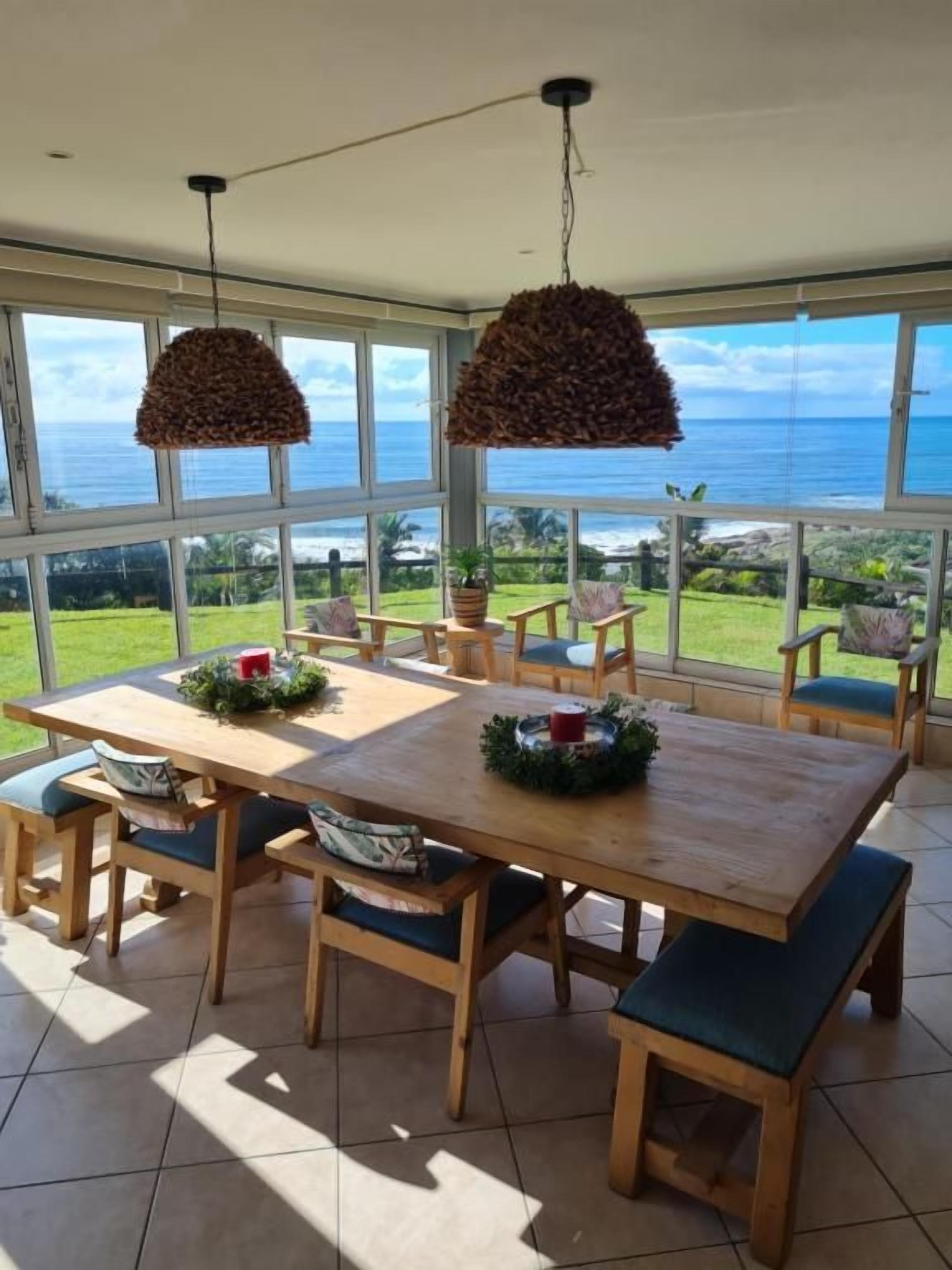A dining area with a large table and chairs, large windows with a view of the ocean, and hanging light fixtures.