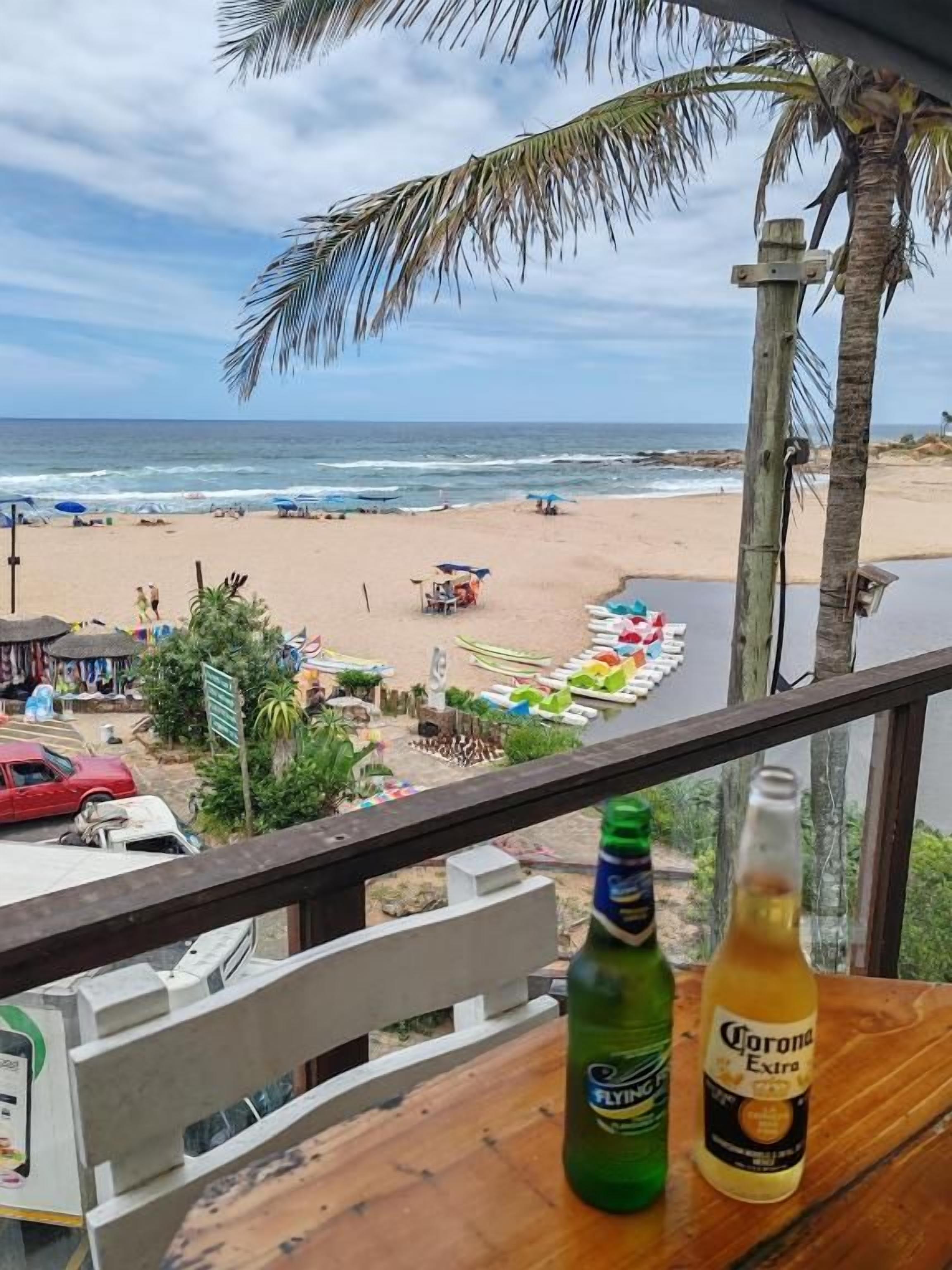Vista al mar desde un balcón con baranda de madera, una botella de cerveza Corona Extra y una botella verde de Flying.