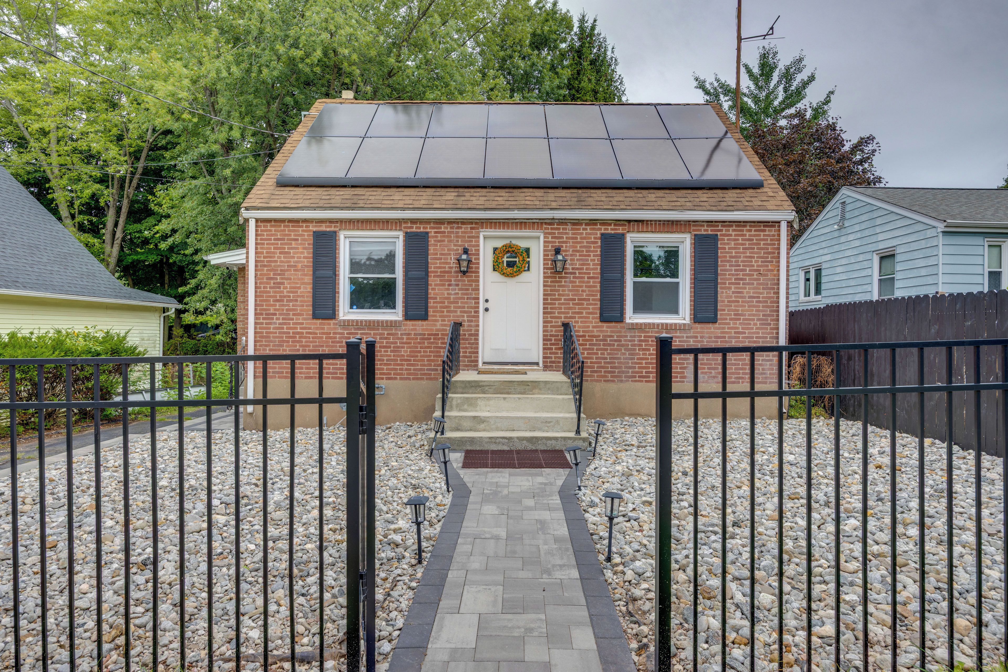 A two-story brick house with a front entrance, a solar panel roof, and a gravel driveway.