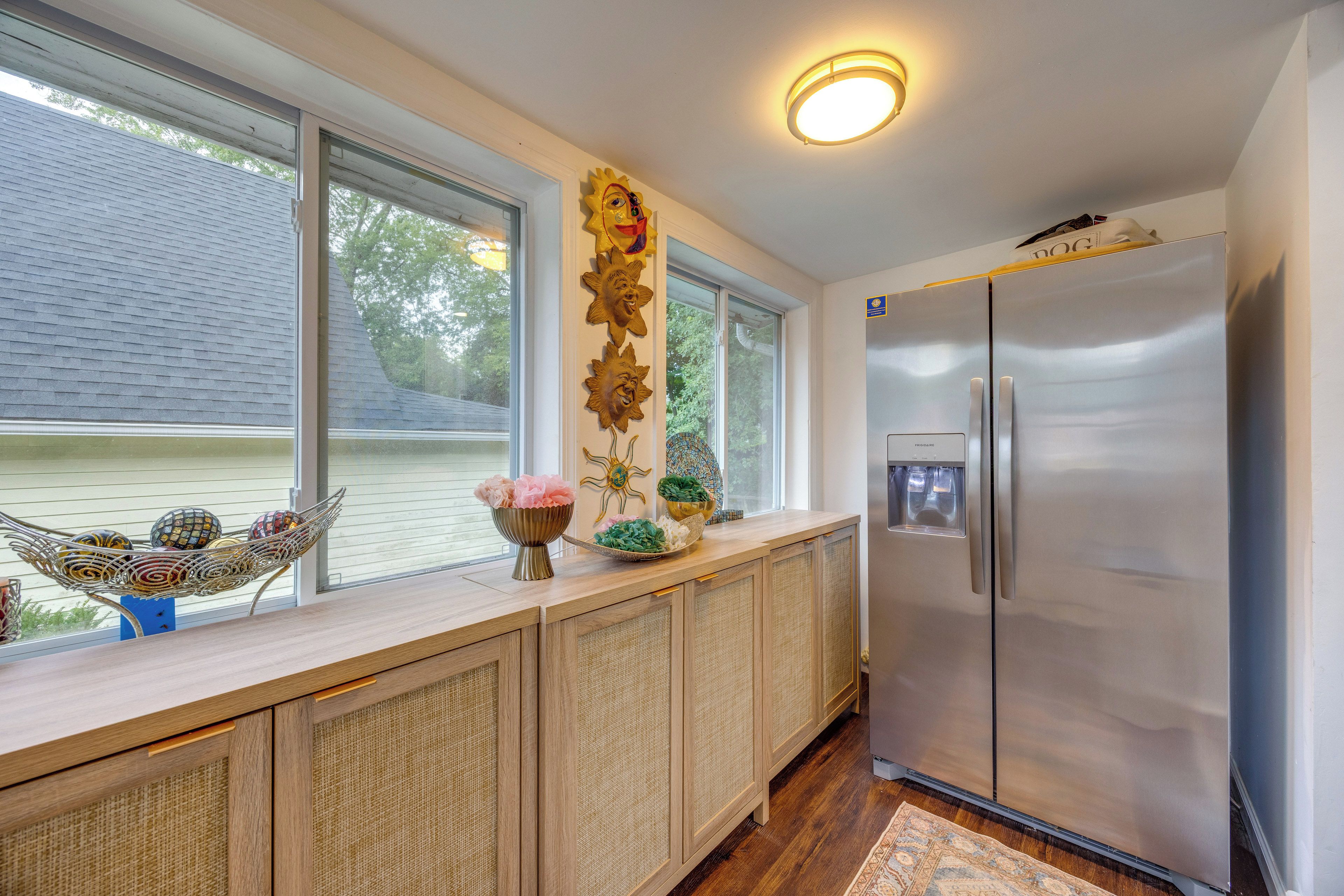 A kitchen with a stainless steel refrigerator, a window with a view of a house, and a decorative wall piece.