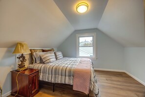 Ett sovrum med ett sluttande tak, en säng med ett quiltat överkast, en sänglampa, ett nattduksbord i trä och ett fönster med utsikt över ett hus.