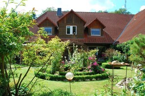 A two-story wooden house with a red-tiled roof, surrounded by a well-maintained garden with various plants and a decorative spherical object.