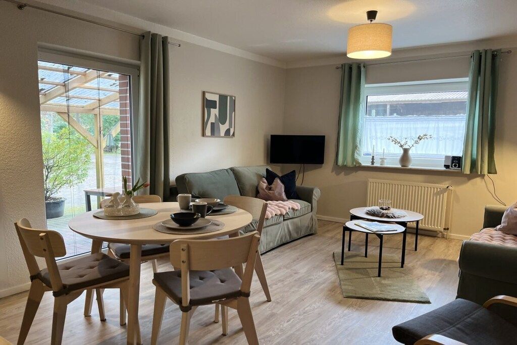 Ein Wohnzimmer mit einer Couch, einem Fernseher, einem Esstisch mit Stühlen und einem Fenster mit Vorhängen.