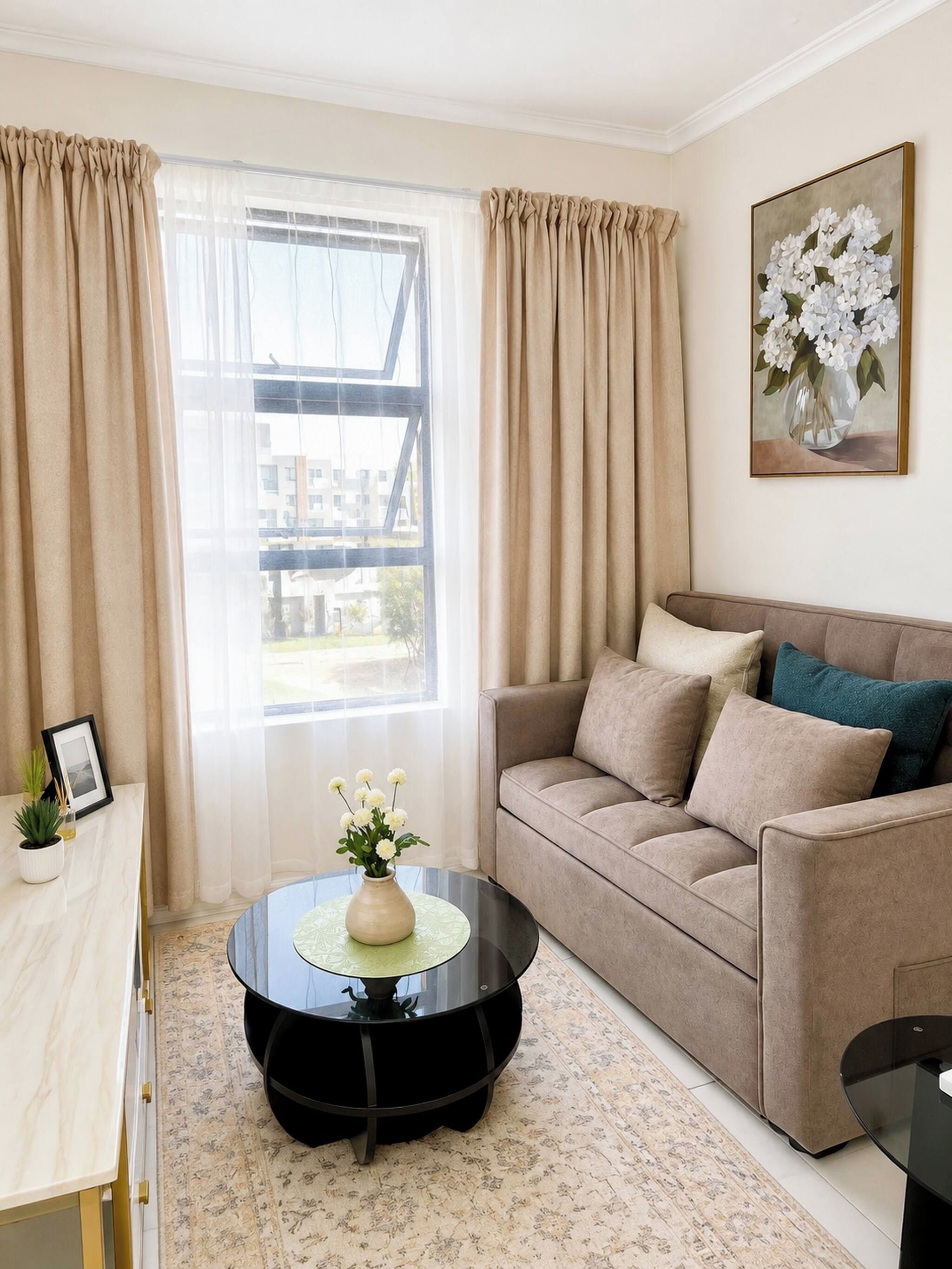 A living room with a beige sofa, a round coffee table with a vase of flowers, and a large window with curtains.