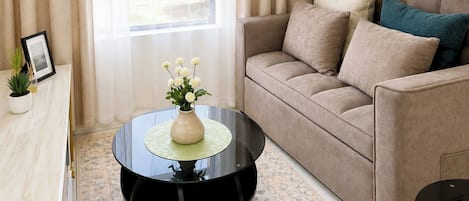 A living room with a beige sofa, a round coffee table with a vase of flowers, and a large window with curtains.