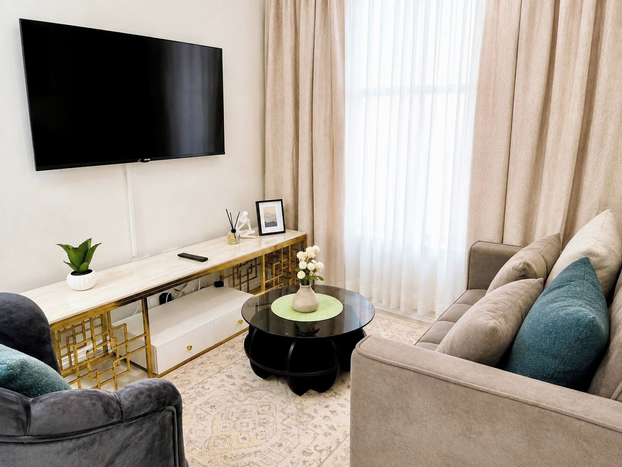 Una sala moderna con un televisor de pantalla plana, una mesa de centro redonda con un florero, un sofá con cojines y un escritorio con una planta y un marco de fotos.