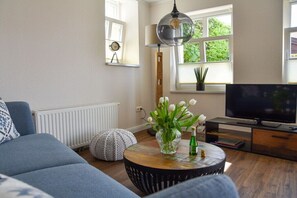 Een woonkamer met een bank, een salontafel met een bloevas, een televisie en een raam met jaloezieën.