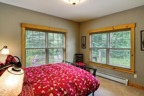 Ein Schlafzimmer mit zwei großen Fenstern, einem Bett mit floralem Überwurf, einem Sessel, einer Lampe und einem gerahmten Bild an der Wand.