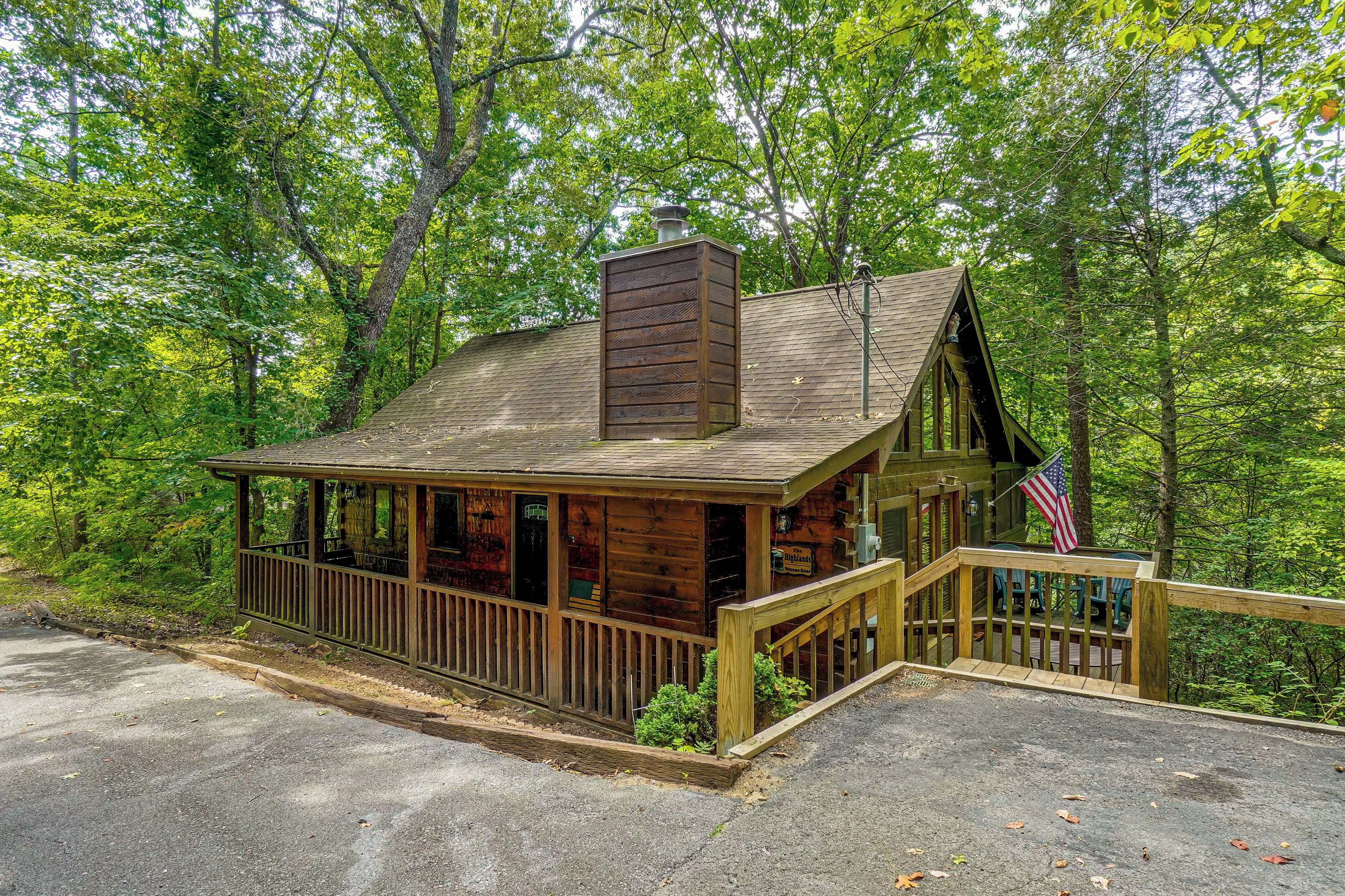 A rustic cabin with a porch and a chimney, surrounded by dense forest.