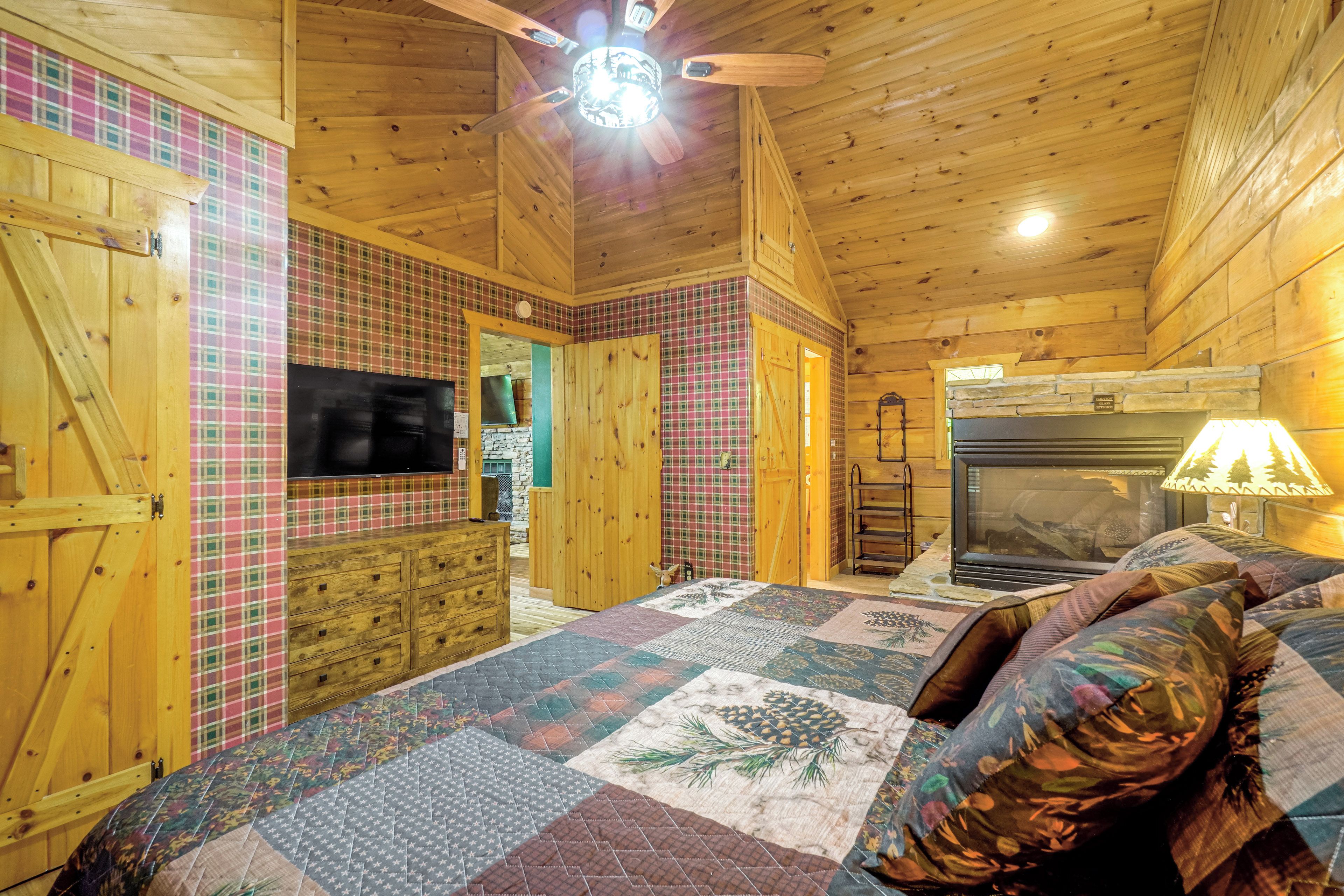 A cozy cabin interior with a wooden ceiling, a fireplace, a bed with a quilt, and a television.