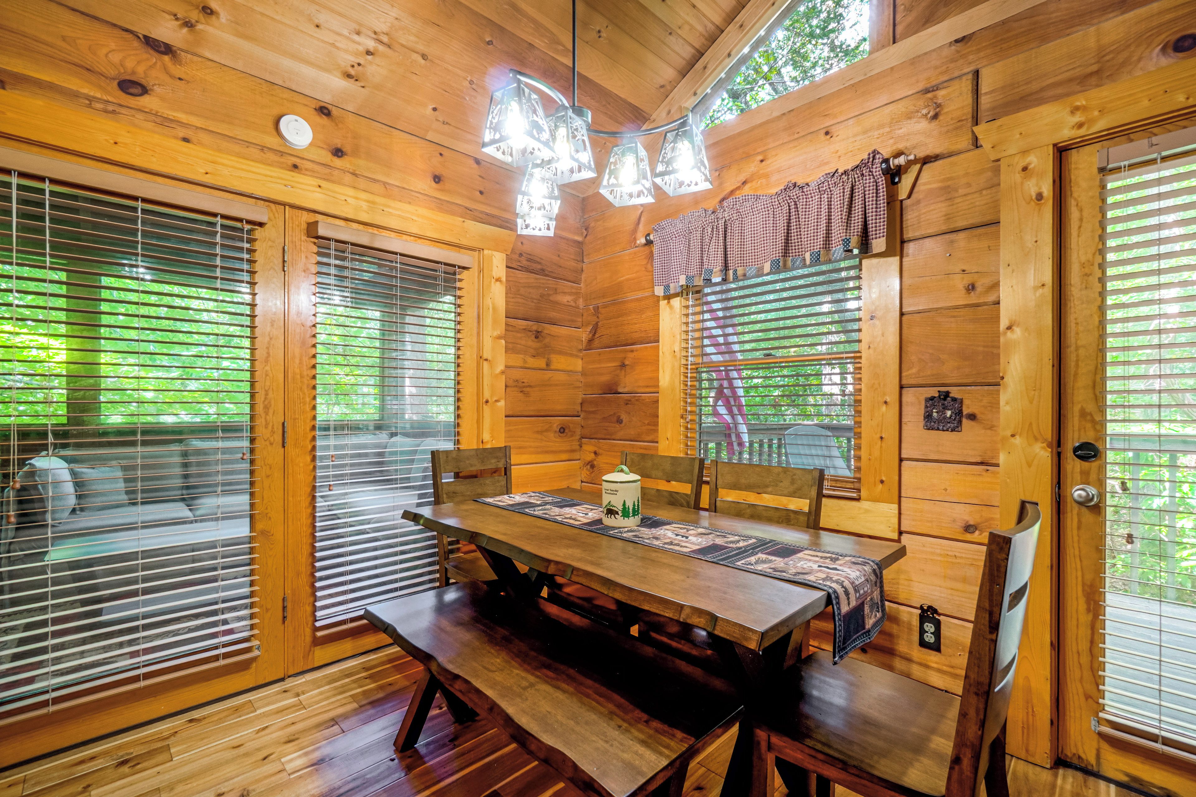 A wooden cabin interior with a dining table, benches, and a window with blinds.