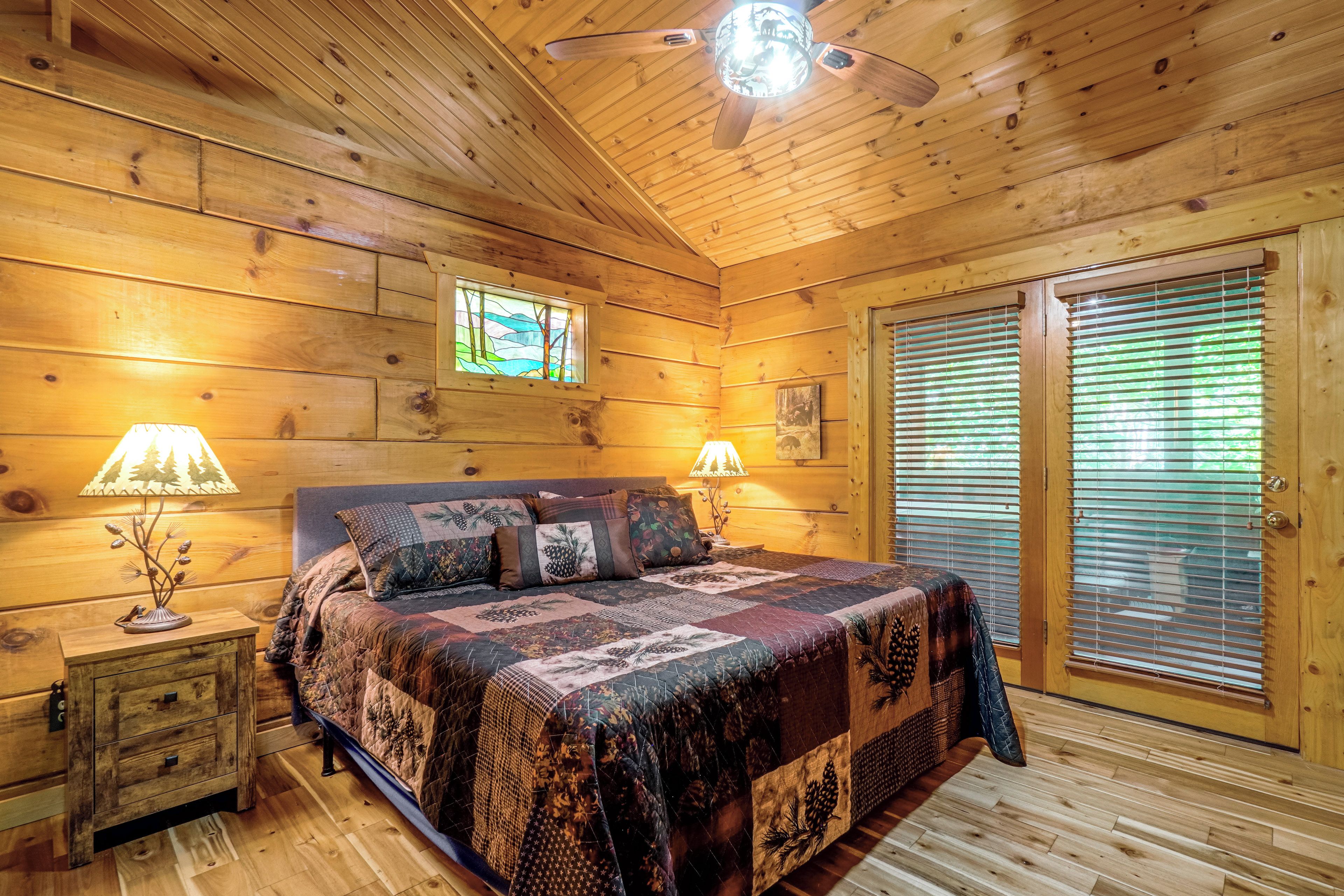 A wooden cabin bedroom with a bed, two bedside lamps, a window, and a door with blinds.