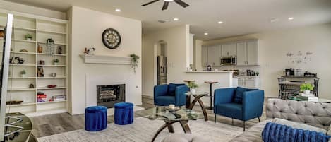Una sala de estar moderna con chimenea, sillones azules, un sofá capitonado y una mesa de centro de vidrio.