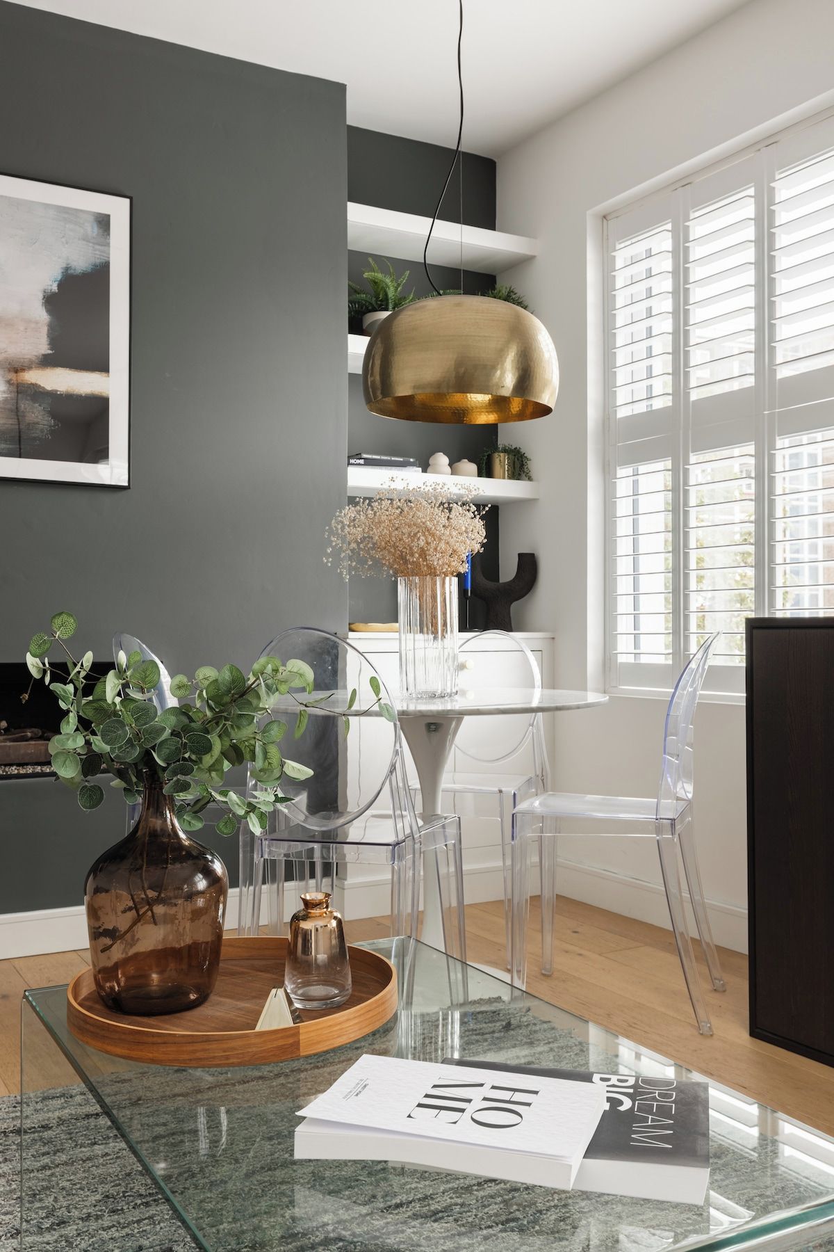 A modern living room with a glass coffee table, a vase with green plants, a gold pendant light, and a view of the city through large windows.