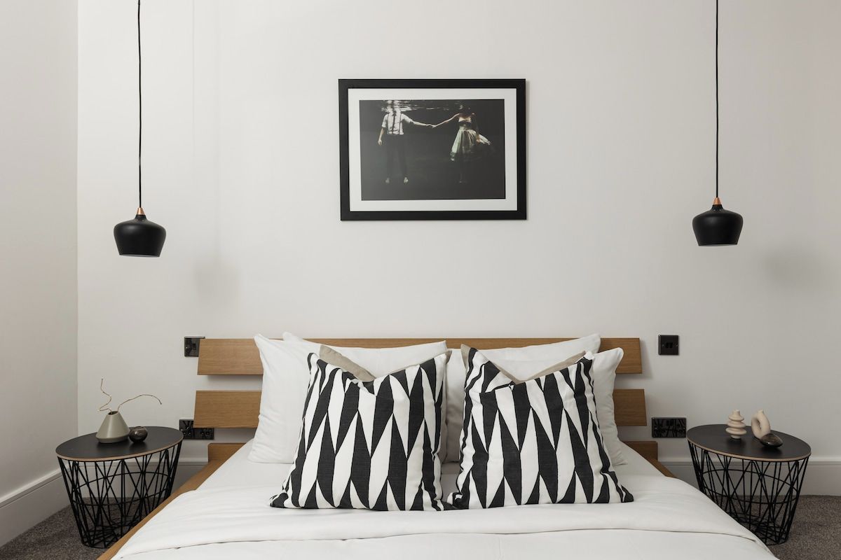 A bedroom with a bed, two nightstands, a framed picture on the wall, and pendant lights.