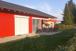 A red building with a garage door, a patio with outdoor furniture, and a tree in the background. - living in a straw house (Gillenfeld)