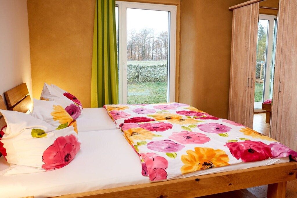 Una cama con colcha y almohadas estampadas con motivos florales, cabecera de madera y vistas a un jardín a través de una ventana.