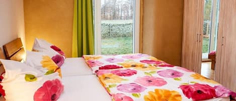 Una cama con colcha y almohadas estampadas con motivos florales, cabecera de madera y vistas a un jardín a través de una ventana.