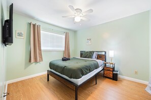 Ein Schlafzimmer mit einem Bett, einem Deckenventilator, einem Fenster mit Jalousien, einem Nachttisch mit Lampe und einem an der Wand befestigten Fernseher.