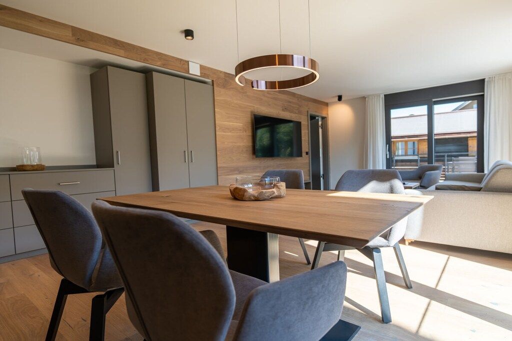 Een moderne eethoek met een houten tafel en stoelen, een aan de muur bevestigde televisie en uitzicht op het buitenleven door een groot raam.