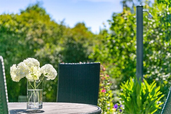 A round table with a glass vase containing white flowers, a dark green chair, and a garden with various plants in the background.